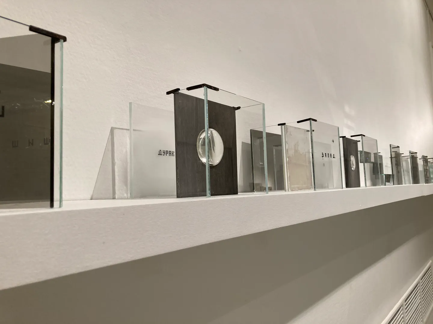 Transparent glass cubes arranged in a row along a wall shelf, each containing translucent plates with Cyrillic text and optical lenses