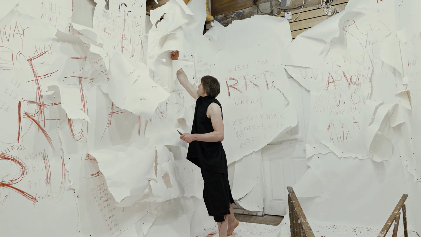 Performance artist reaching up against gallery walls covered with layered torn white paper sheets and handwritten red text