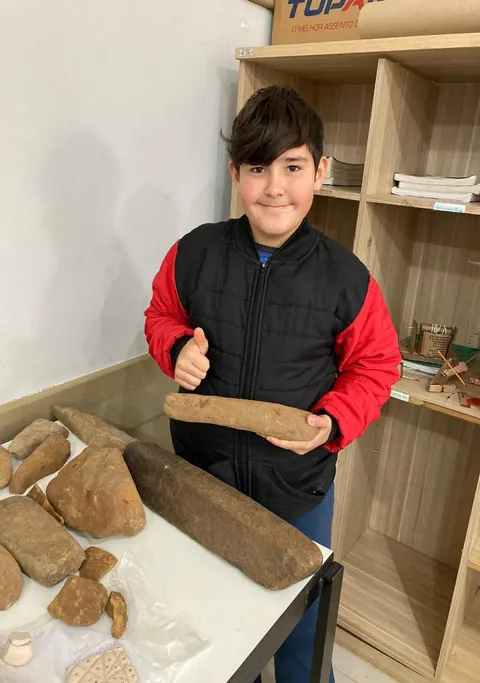 O aluno Vitor Hugo Bortolini. 6º ano encontrou várias peças arqueológicas na propriedade de sua família, localizada na comunidade de Irakin. No interior do município de Tangará. O artefato que ele mostra na imagem, uma rocha ígnea, é uma mão de pilão. Peso: 2,900kg. Comprimento:31cm. Largura: 10cm. Espessura:4,5cm.