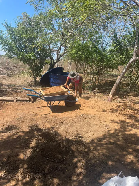 Worker using a wooden sieve frame to sift soil into a wheelbarrow near a water reservoir tank.