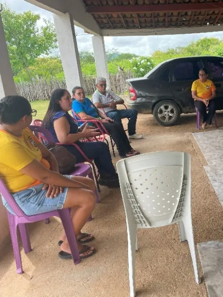 Attendees and project coordinators seated in chairs under a covered porch during a community meeting.